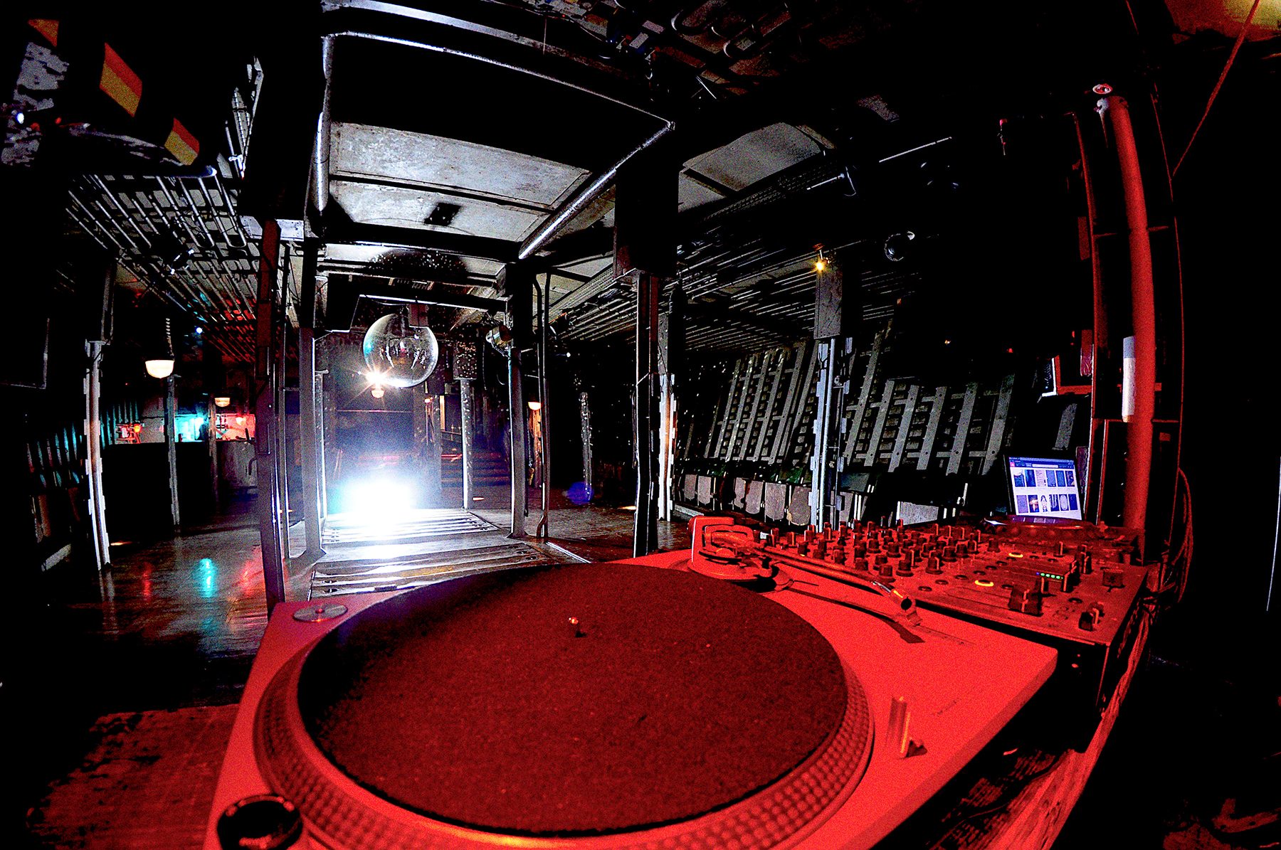 Ein Turntable in rotem Licht fotografiert, mit dem leeren Laderaum 1 im Hintergrund. In der Mitte des Raums hängt eine Discokugel, die von einem weißen Scheinwerfer angestrahlt wird.