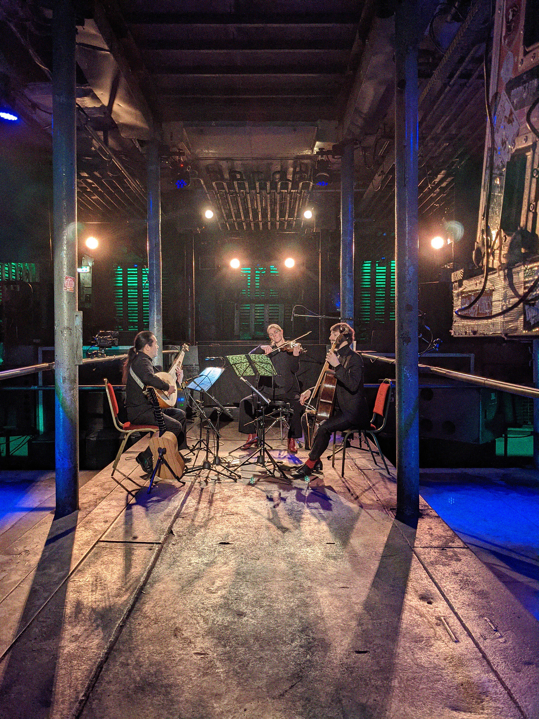 Ein Konzert auf dem Wellentunnel, vor der Bühne in Laderaum 4. Drei Menschen sitzen im Kreis und spielen Instrumente auf der hell beleuchteten Tanzfläche von Laderaum 4.