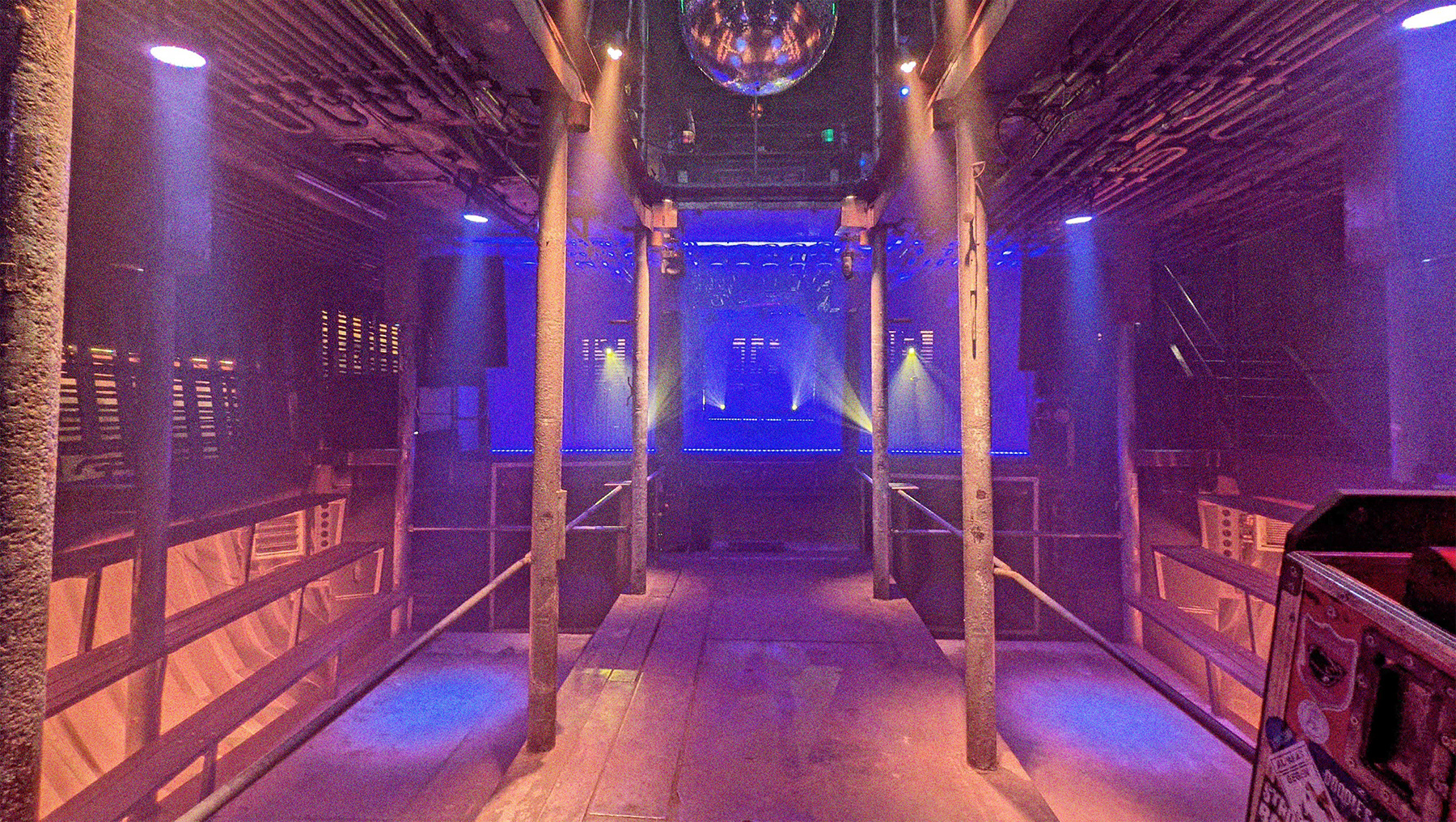Der leere Laderaum 4 mit Blick frontal auf die Bühne. Die Tanzfläche befindet sich in der Mitte. An den Seiten links und rechts befinden sich Sitzbänke aus Holz. Scheinwerfer beleuchten den Raum in blau und lila.