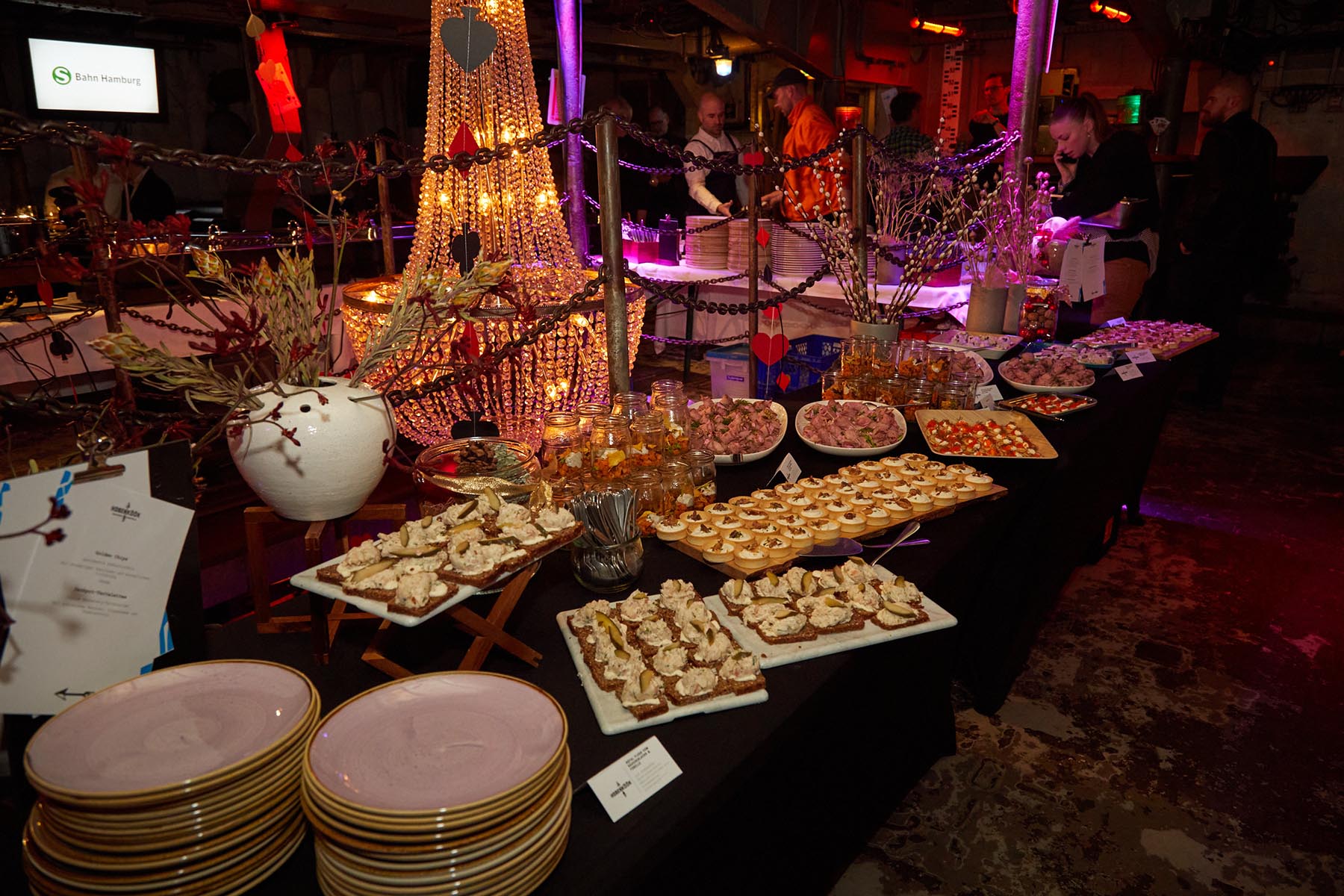 Buffet mit verschiedenen Gerichten in Gläsern, Platten und links hängt ein Kronleuchter. Der Raum ist warm, lila beleuchtet. 