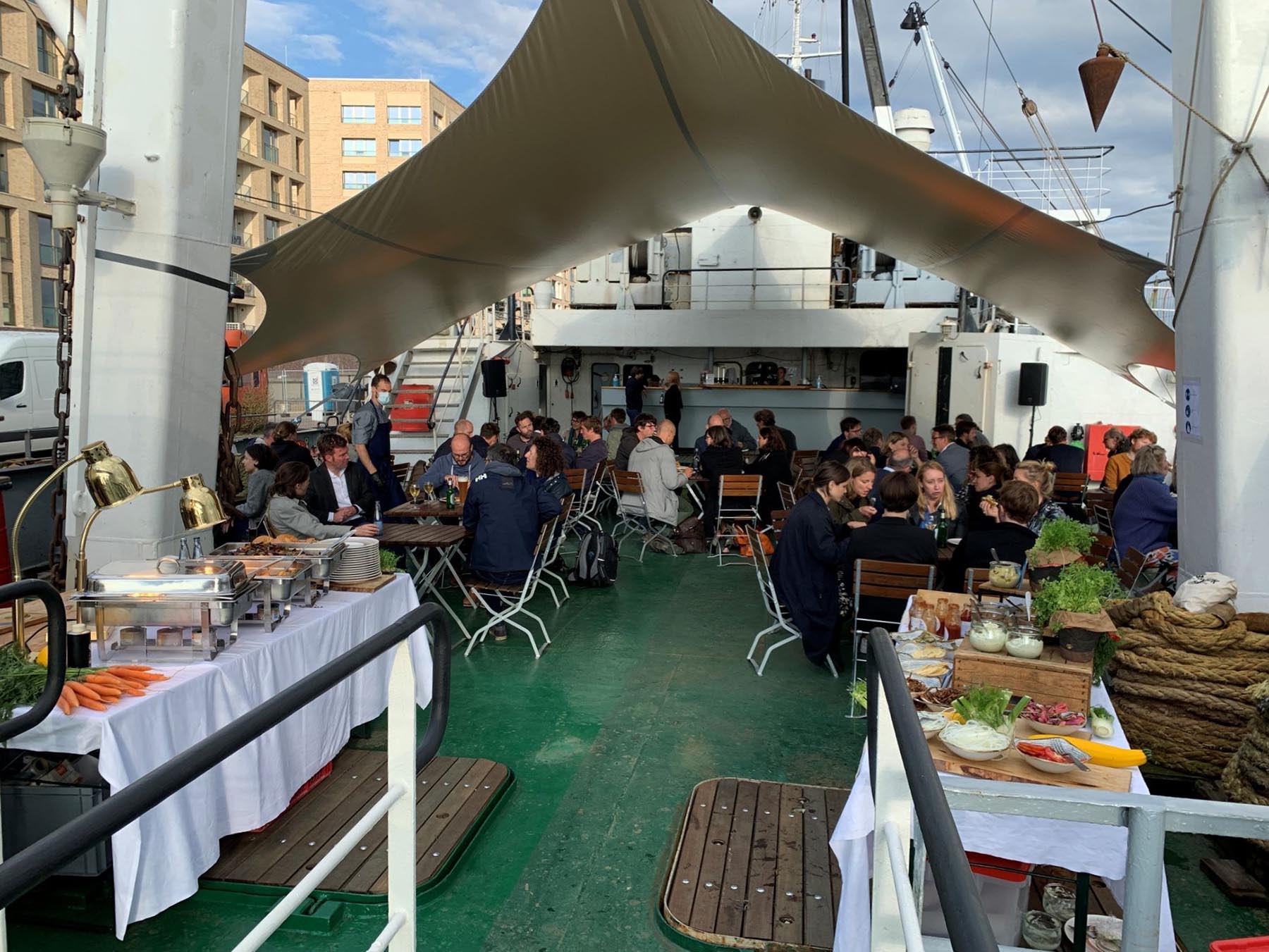 Buffetaufbau auf dem Achter Deck der Stubnitz. Gäste sitzen unter einem Sonnensegel zum Essen im Freien.