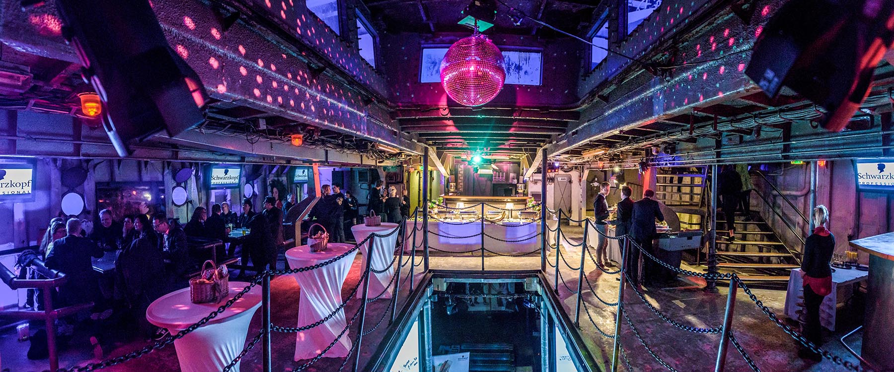 Event an Bord der MS Stubnitz: Stehtische, ein Buffet, links sitzen Menschen, in der Mitte des Raumes hängt eine Discokugel.