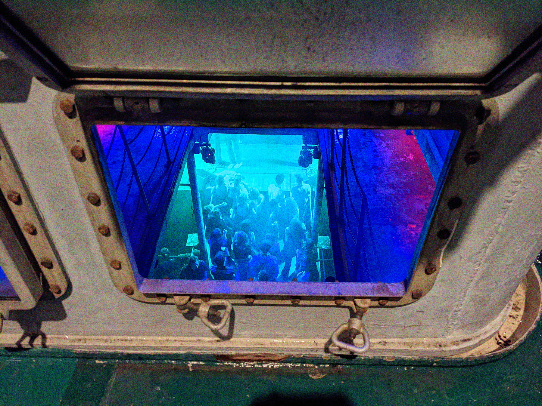 Der Blick auf die Tanzfläche in Laderaum 4, fotografiert durch die Ladeluke auf dem Achterdeck. Blaues Licht im Inneren des Schiffes. Unten tanzen Menschen.